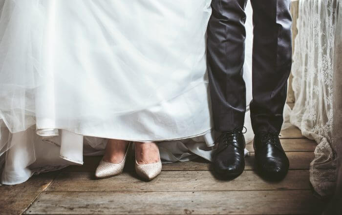 Close-up of a couple's lower legs on a wooden floor. The woman wears a white dress and sparkly heels, while the man wears dark pants and black shoes.