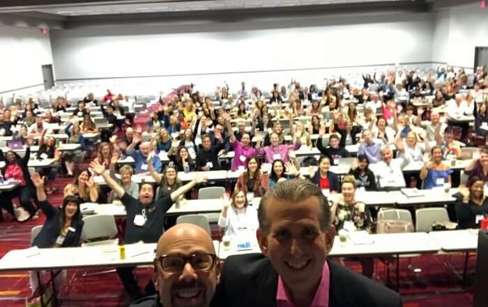 Two people take a selfie in front of a large group seated in a conference room. The audience waves and smiles.