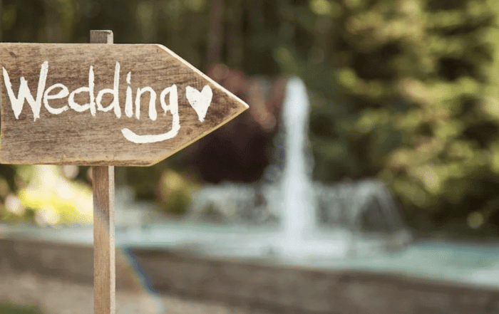 Wooden sign with the word "Wedding" and a heart symbol, pointing right. A blurred garden and fountain are in the background.
