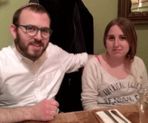 A man and a woman sit together at a table in a restaurant. The man wears glasses and a white shirt, and the woman wears a light-colored top. A coat hangs in the background.