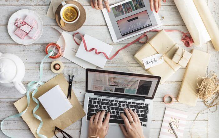 Two people work at a table. One uses a laptop, and the other holds a tablet. The table is covered with gift wrapping materials, a cup of tea, and two pink pastries.