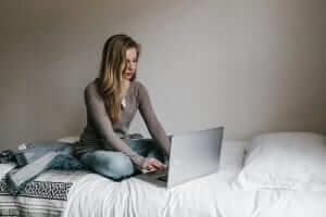 A person sits on a bed using a laptop, with a jacket and a blanket beside them.