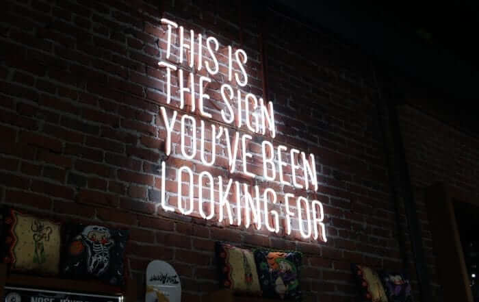 Neon sign on a brick wall reads, "This is the sign you've been looking for" with decorative pillows below.