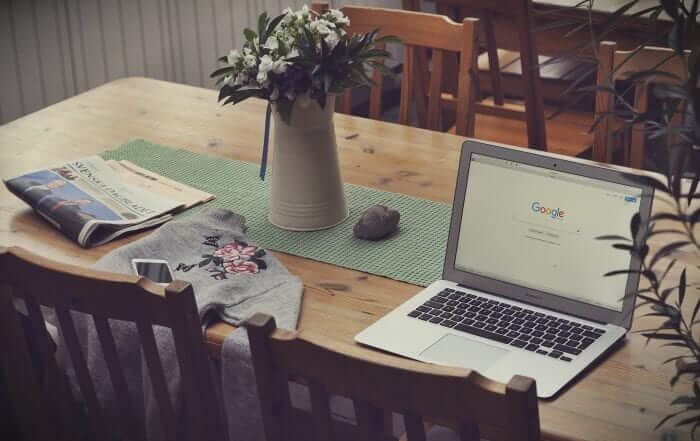 A wooden table with a laptop open to Google's homepage, a vase of flowers, a newspaper, a sweatshirt, and a smartphone.