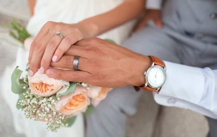 Close-up of a couple holding hands over a bouquet of pink roses. The woman has a diamond ring and the man wears a watch and wedding band.