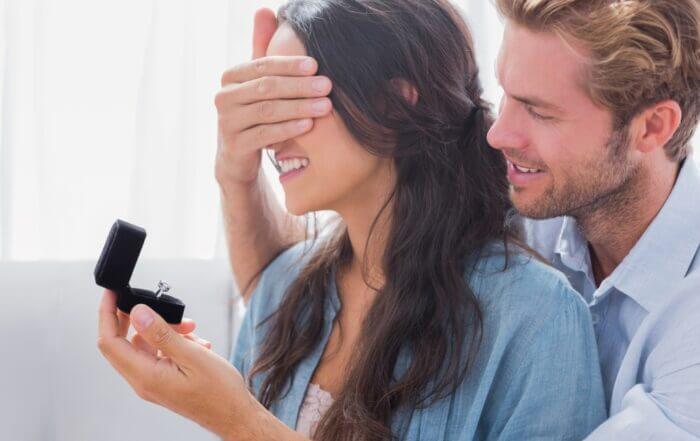 A man covers a woman's eyes while holding an open ring box with an engagement ring.