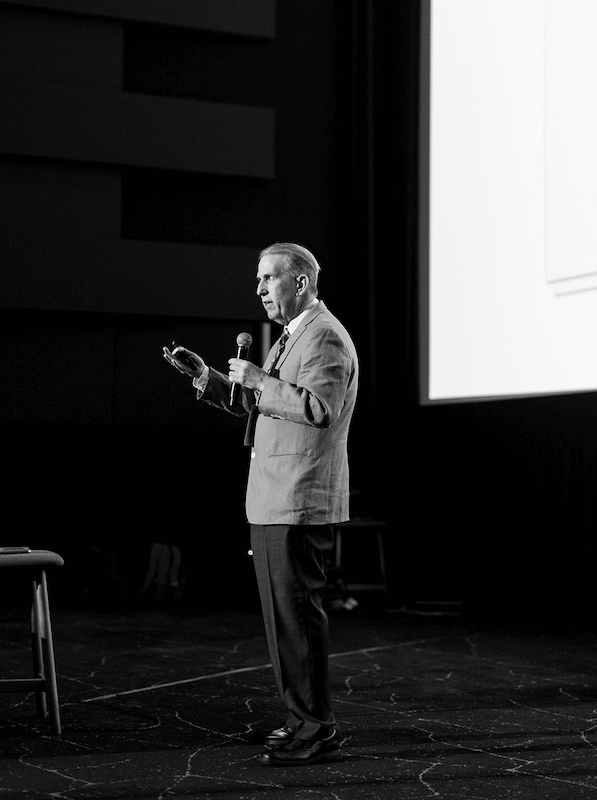 keynote speaker Brian Lawrence A person in a suit gives a presentation on stage, holding a microphone and a clicker, with a projection screen in the background.