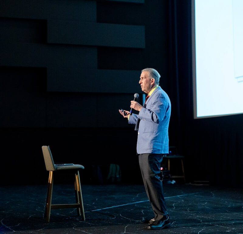 DJ Collective 2023 Person in a blue jacket speaking into a microphone on stage, with a chair beside them and a large screen in the background.