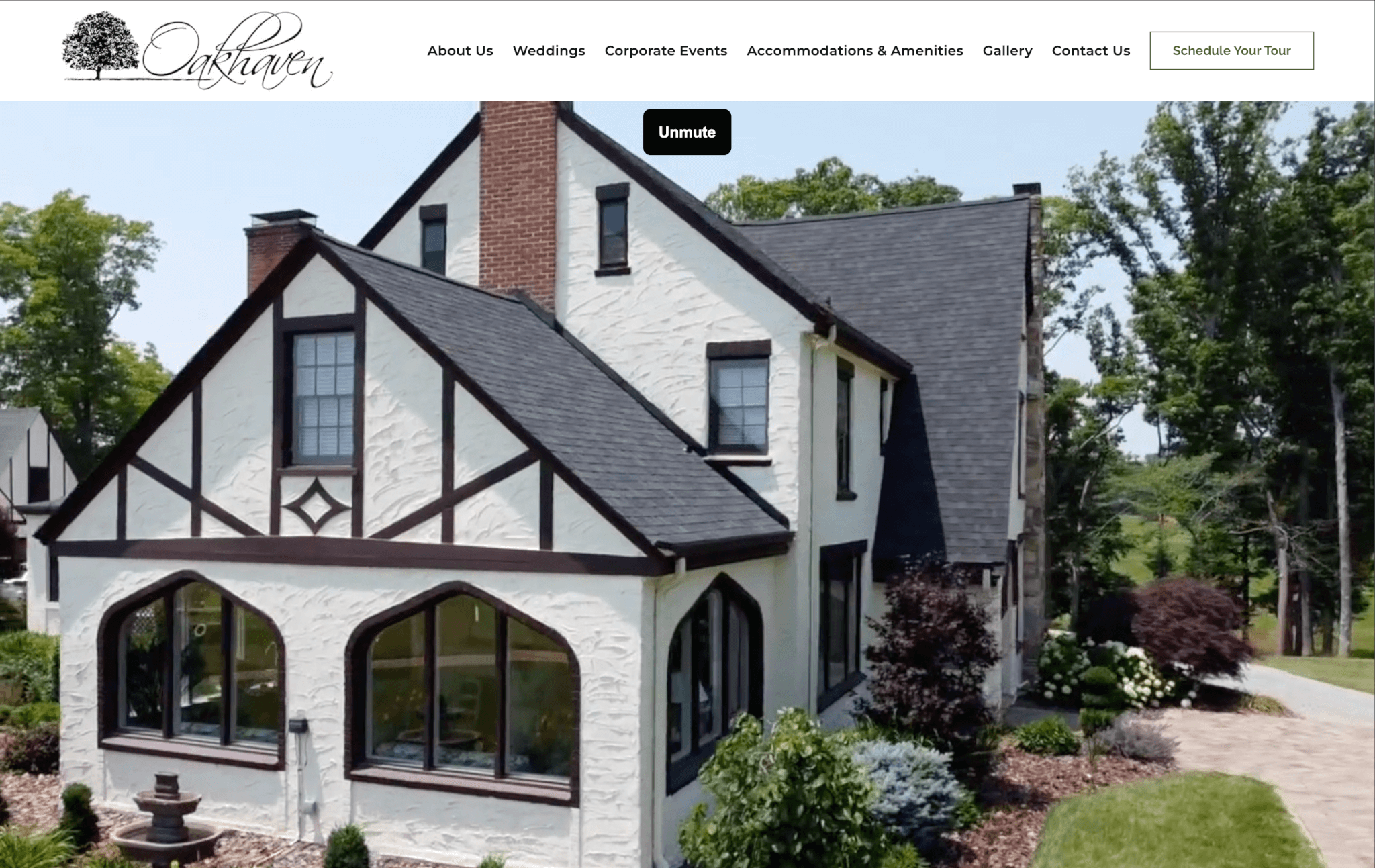 A two-story Tudor-style house with white walls and dark beams, surrounded by trees and manicured bushes, under a clear sky.